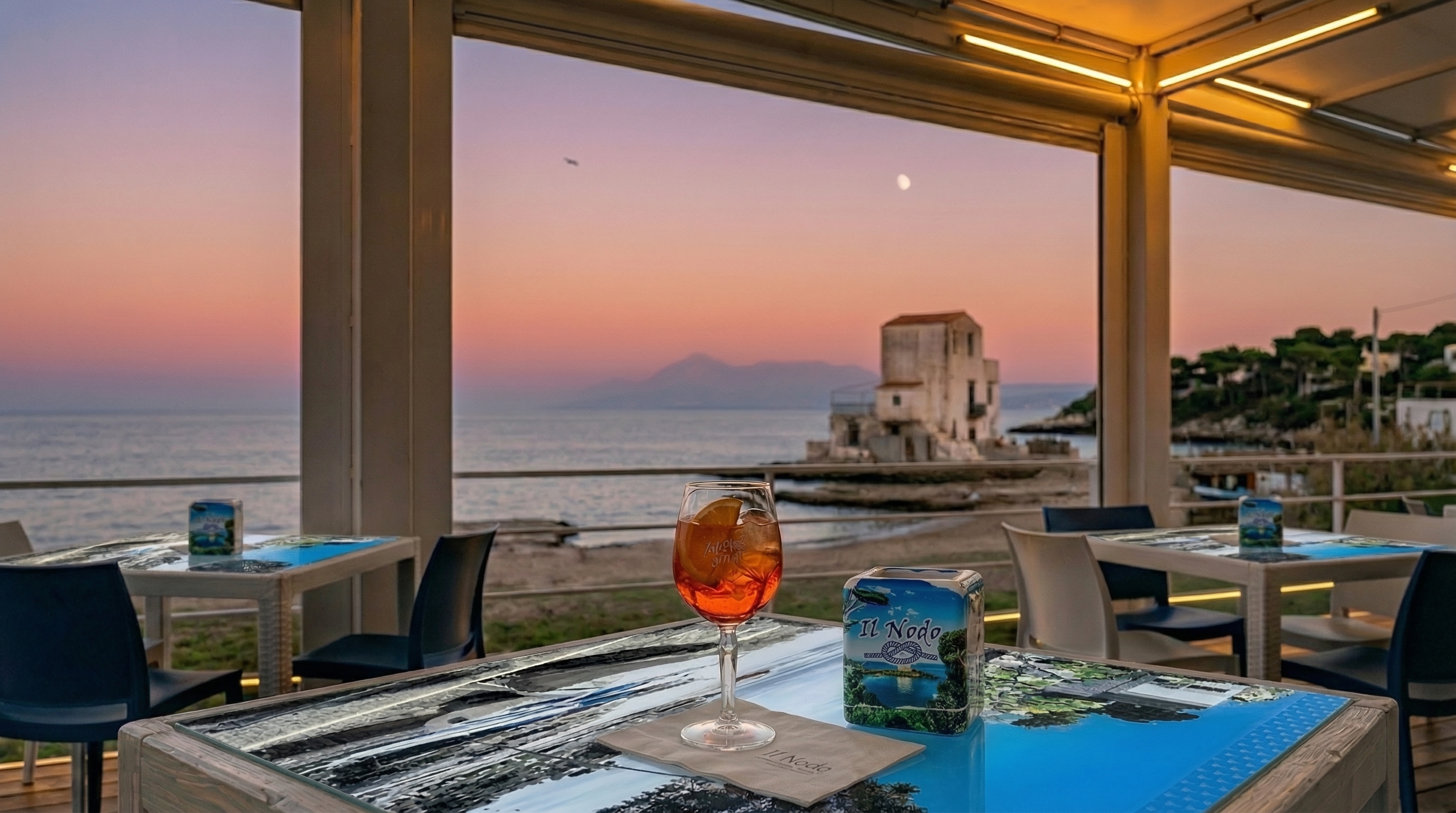 Vista panoramica del beach bar Il Nodo 2.0 a Sant'Elia con la celebre casa bianca sugli scogli e un Aperol Spritz sul bancone